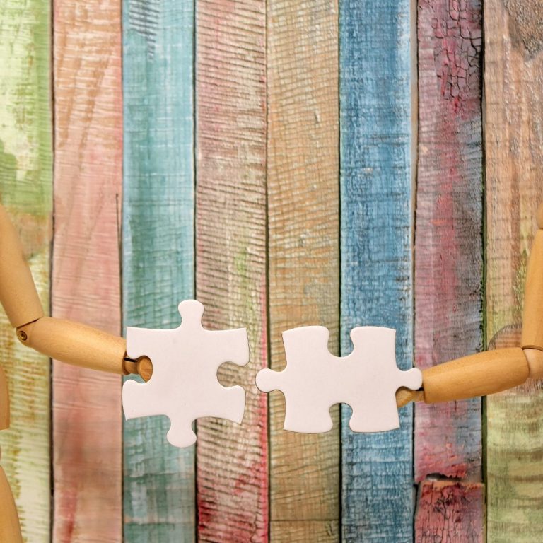 Puzzle Zwei Holzfiguren halten jeweils ein Puzzlestück vor einem bunten Holzhintergrund.